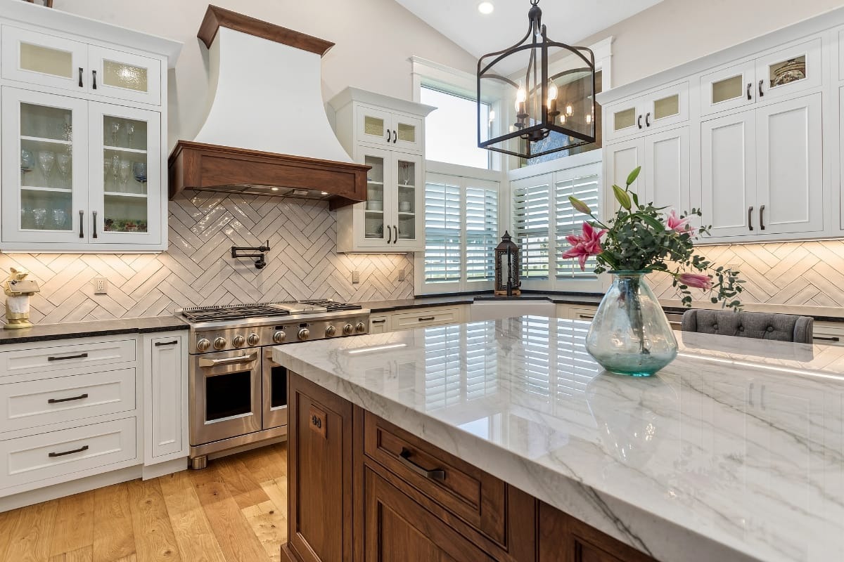 Modern kitchen with white cabinets, marble island countertop, stainless steel stove, wood accents, herringbone backsplash, and a vase of flowers on the island.