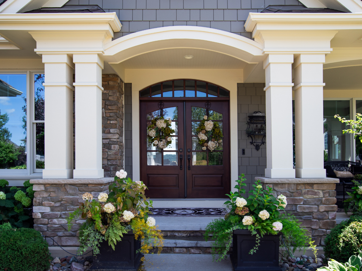 Front entrance of a house with double wooden doors—a popular choice among door types—adorned with floral wreaths, stone and siding exterior, potted plants, and decorative columns.
