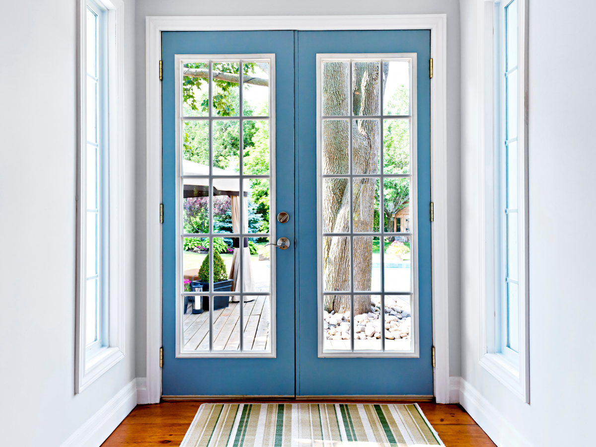 Double glass French doors—a classic among door types—with blue frames open to a backyard featuring a tree, patio furniture, and a swimming pool. A striped rug adorns the wooden floor just inside the entryway.