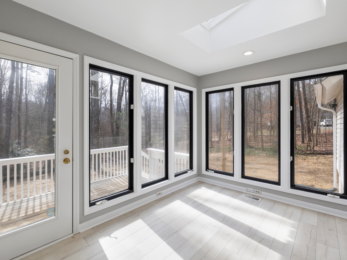 Bright sunroom with large windows, light wood flooring, a skylight, and a stylish door—one of several door types available—leading to a white-railed deck. Forested area visible outside.