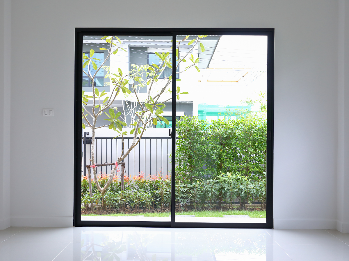 Large glass sliding door, one of the most popular door types, with a black frame opens to a garden with green shrubs, a small tree, and a black metal fence in a modern residential area.