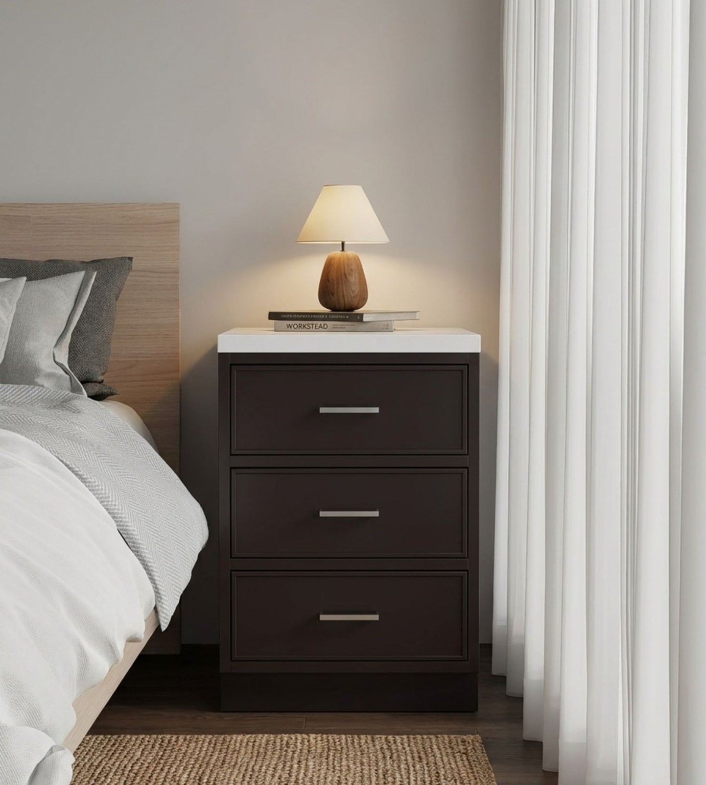 A nightstand with three drawers sits beside a bed, topped with a closed book and a small lamp—an inviting contrast to the sleek lines of an office bathroom remodel. White curtains cover the window to the right.