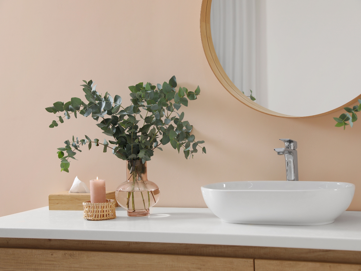 Modern bathroom vanity featuring a white sink, faucet, glass vase of eucalyptus branches, round mirror, tissue box, and a pink candle on a wicker tray.