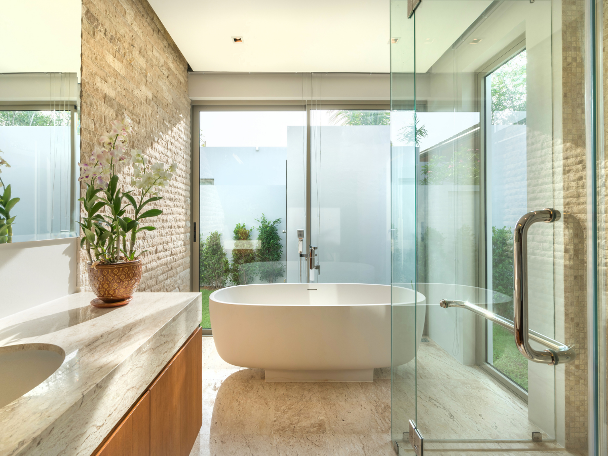Modern bathroom with a freestanding white bathtub, glass shower enclosure, stylish bathroom vanity featuring a stone countertop with sink, potted plants, and large frosted windows overlooking a small garden.