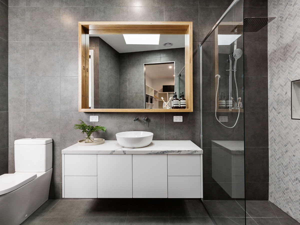 Modern bathroom with a large mirror, bathroom vanity featuring a vessel sink, potted plant, toiletries, toilet, and a glass-enclosed shower with a rainfall showerhead.
