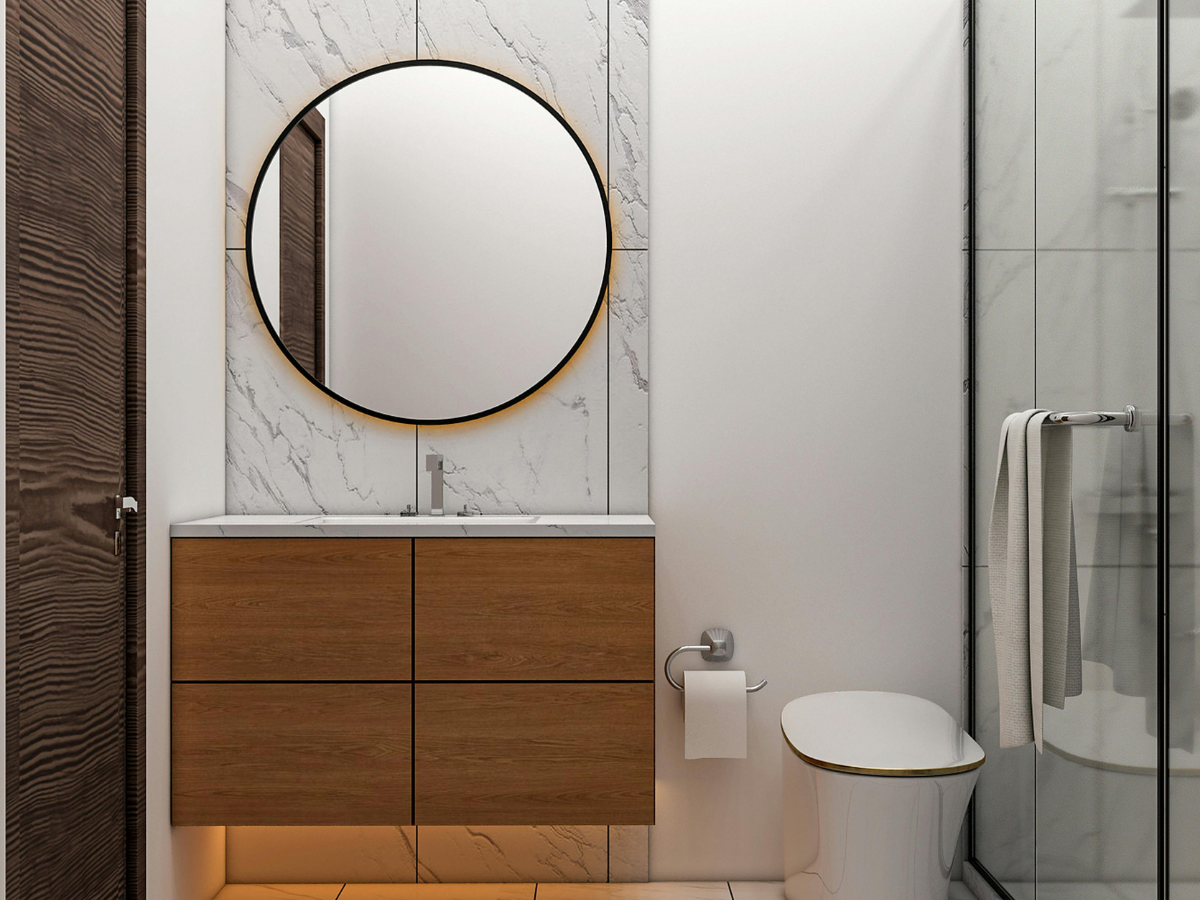 Modern bathroom with a round mirror, sleek bathroom vanity, wall-mounted faucet, toilet, toilet paper holder, and a glass shower with white marble tiles and a towel hanging inside.