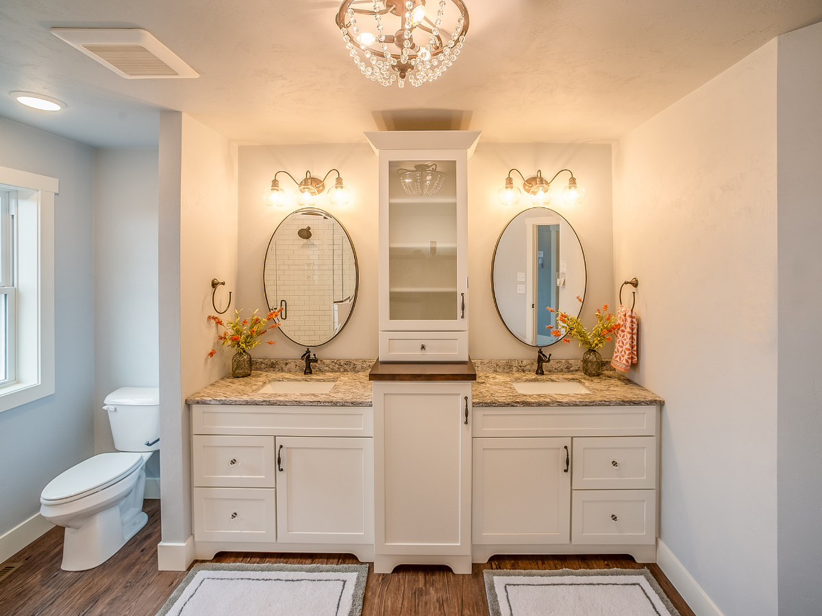 Bright bathroom with double sinks set in a sleek bathroom vanity, round mirrors, white cabinets, granite countertop, floral arrangements, and toilet on the left; overhead lighting and wood-style flooring.