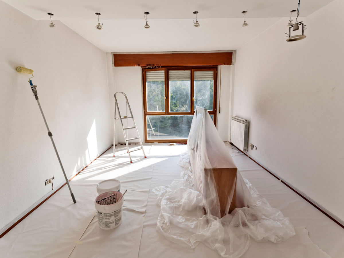A room prepared for painting with plastic sheets covering the floor and furniture, a ladder, paint roller, and paint cans visible—an ideal setting to consult a paint sheen guide before getting started.