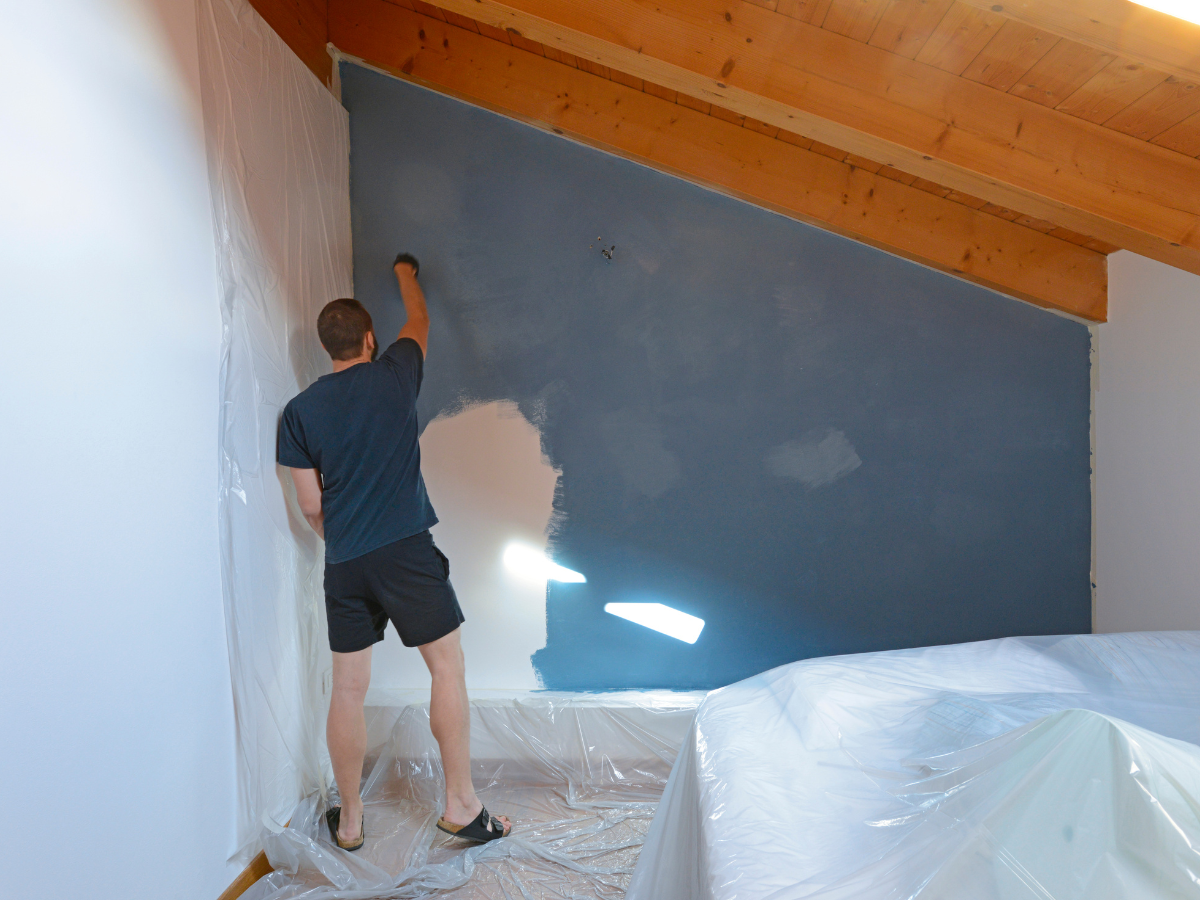 A person in shorts and a t-shirt paints a slanted wall blue in a room with a wooden ceiling. Plastic sheets cover the floor and furniture for protection, while a nearby paint sheen guide helps ensure the perfect finish for the freshly painted surface.