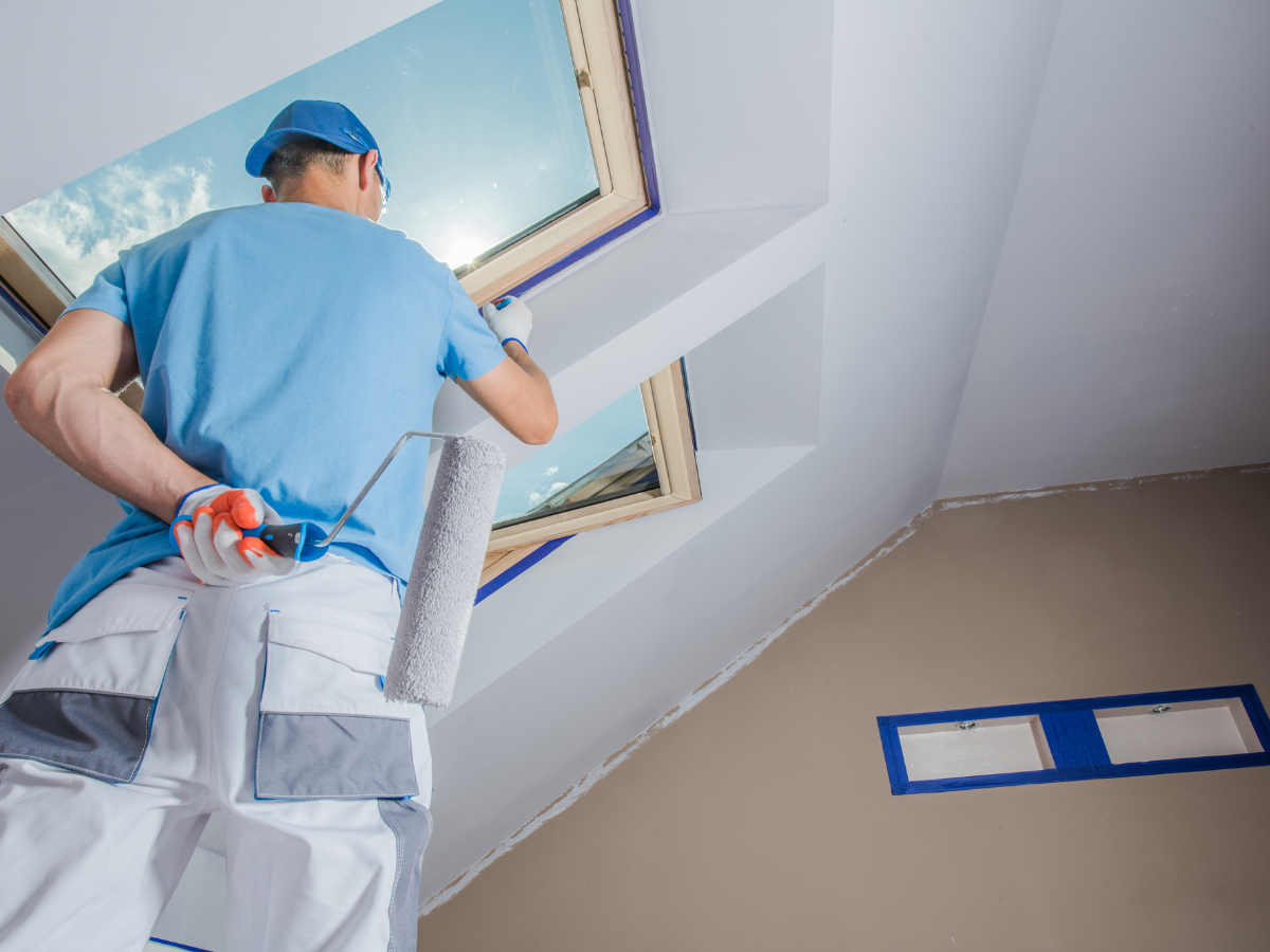 Person in work clothes paints a slanted ceiling near a skylight window, holding a paint roller and wearing a blue cap. Blue tape covers window edges for protection, ensuring clean lines while following a paint sheen guide for the perfect finish.