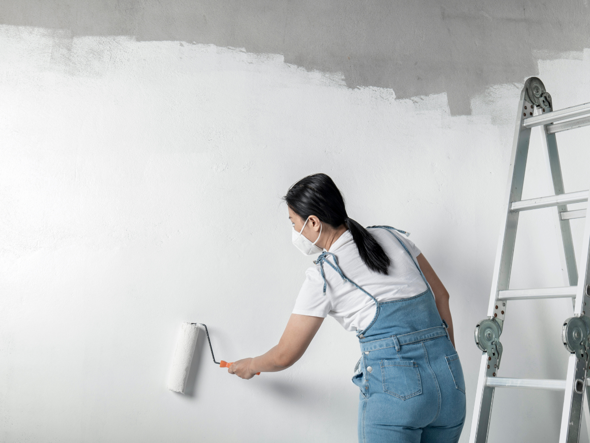 A woman wearing a mask and overalls paints a wall white with a roller, using techniques from a paint sheen guide. A ladder stands nearby.