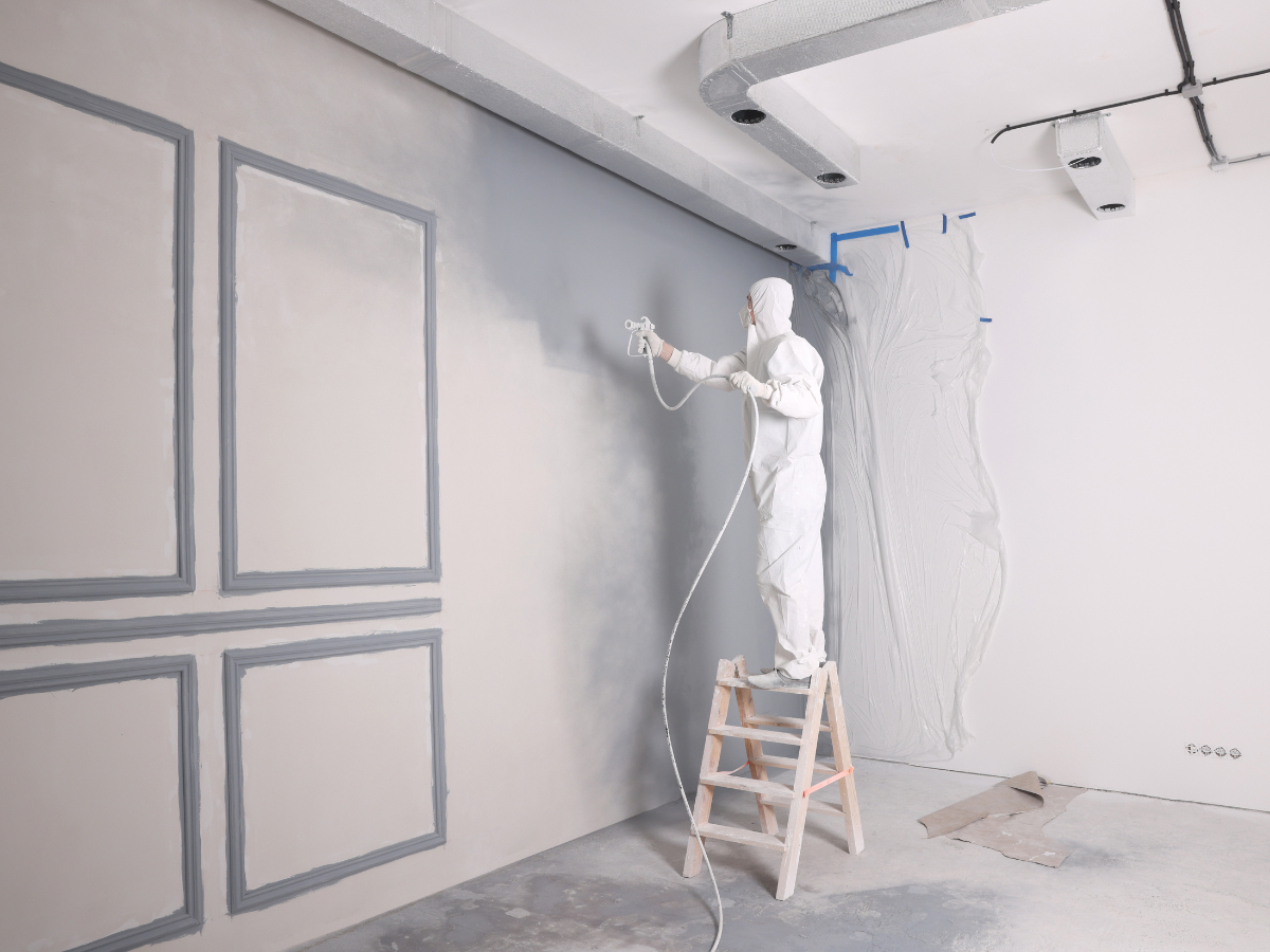 Person in protective suit stands on ladder spray-painting a large wall in a room under renovation; some areas are masked with plastic, following a detailed paint sheen guide to ensure the perfect finish.