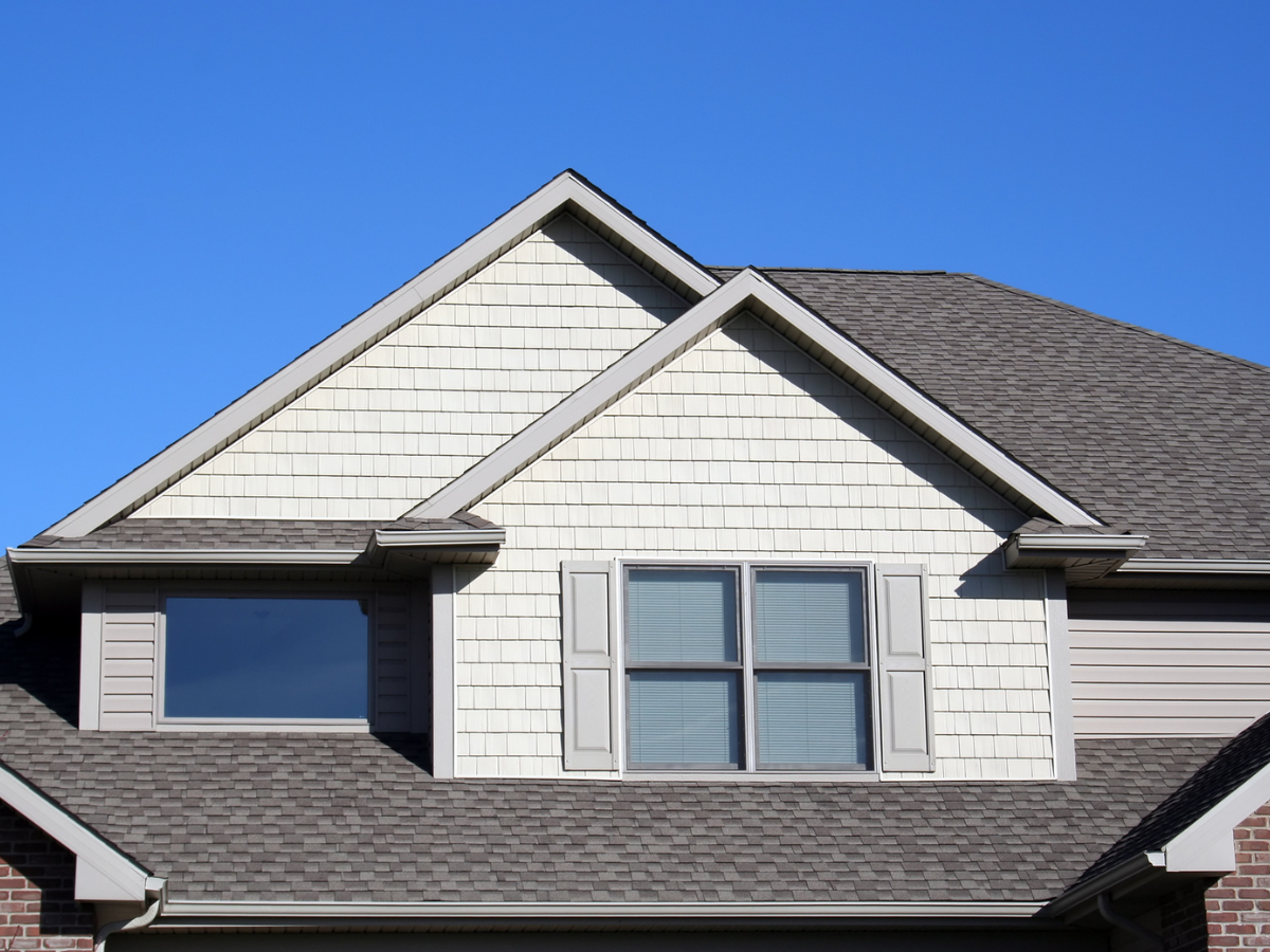 Close-up of a house exterior shows gray roof shingles made from durable roofing materials, white siding, two upper windows, and a clear blue sky in the background.