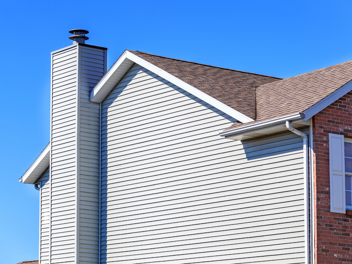 Side view of a house exterior with beige vinyl siding, a brick wall section, a chimney, and a brown shingled roof made from quality roofing materials against a clear blue sky.