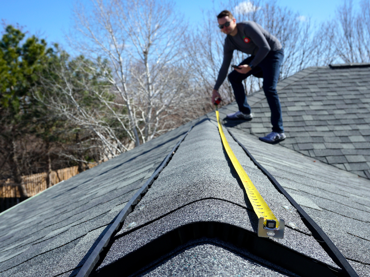 A person measures the length of a shingled roof ridge with a yellow tape measure on a clear, sunny day, carefully assessing the area to determine the amount of roofing materials needed.