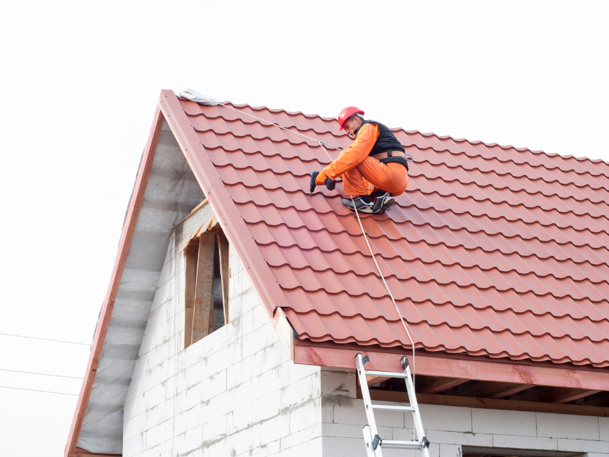 Worker in orange safety gear and helmet attaches a tether while working on the red metal roof of a building under construction, securing himself among the roofing materials, with a ladder leaning against the roof edge.
