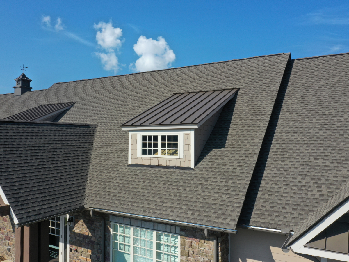 A close-up view of a house roof with gray shingles showcases quality roofing materials, featuring a dormer window and a clear blue sky in the background.