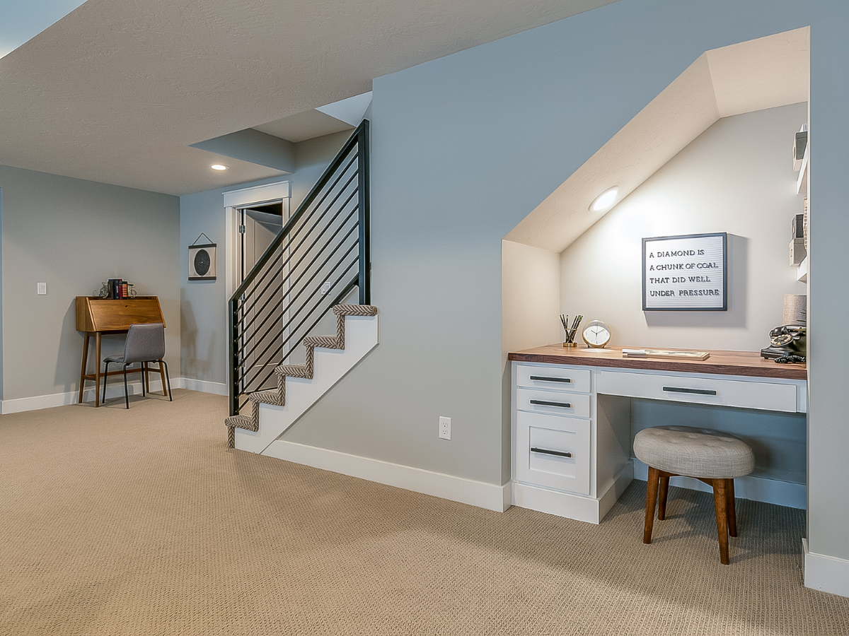 Modern basement with a carpeted floor, staircase with metal railings, under-stairs desk area, stool, and wall art. Showcasing thoughtful basement office design, a second desk with chair is in the background.