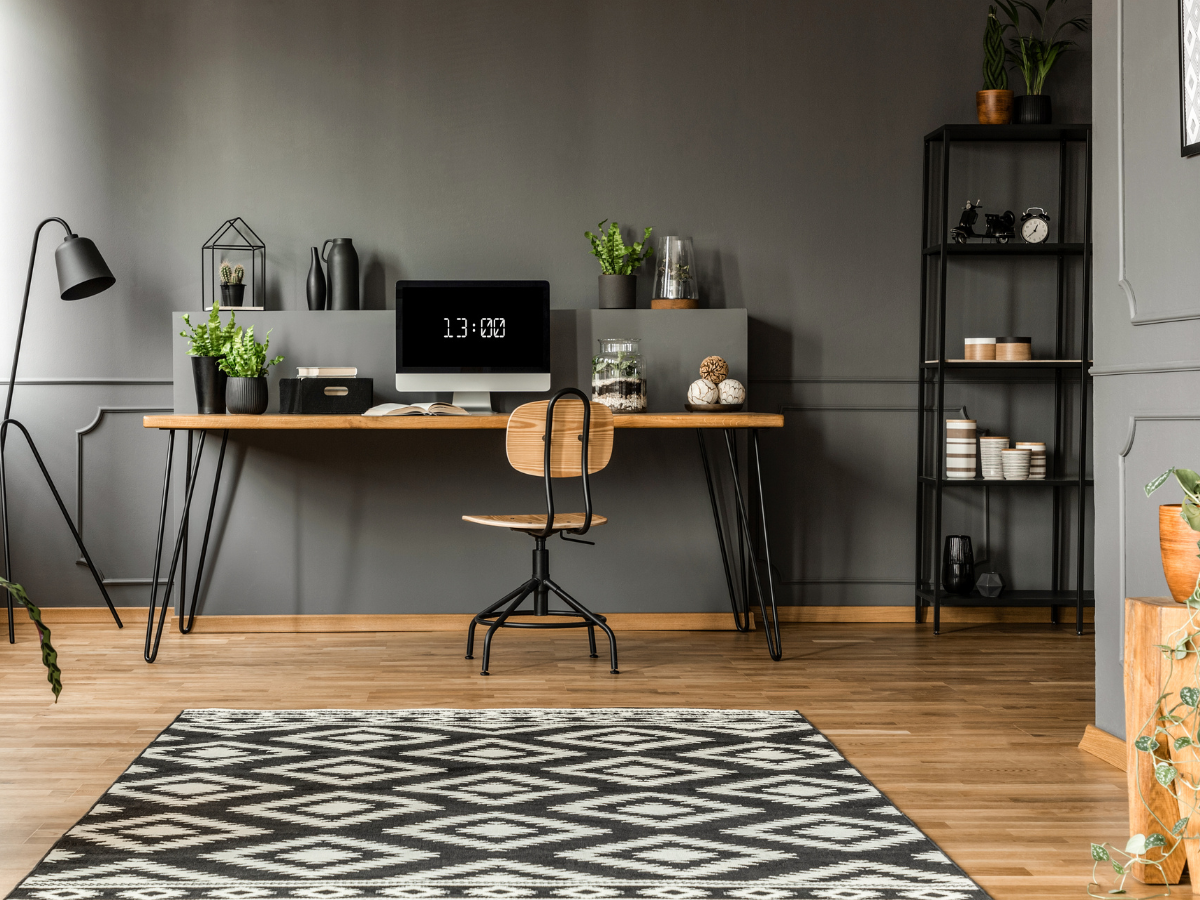 Modern basement office design featuring a wooden desk, computer, chair, plants, shelves, black lamp, and a geometric patterned rug on a wooden floor.
