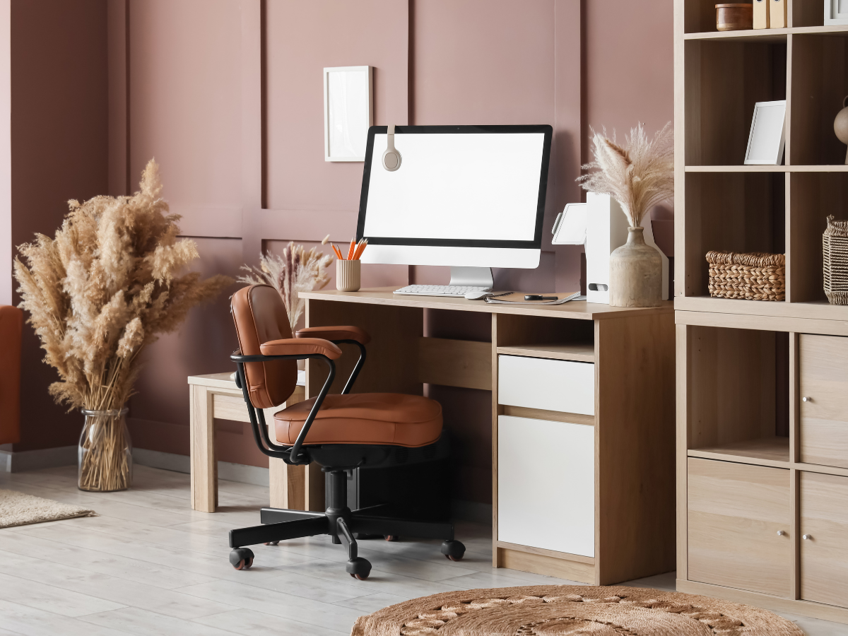 A modern basement office design features a computer on a wooden desk, a leather office chair, pampas grass decor, and shelves against a mauve wall.