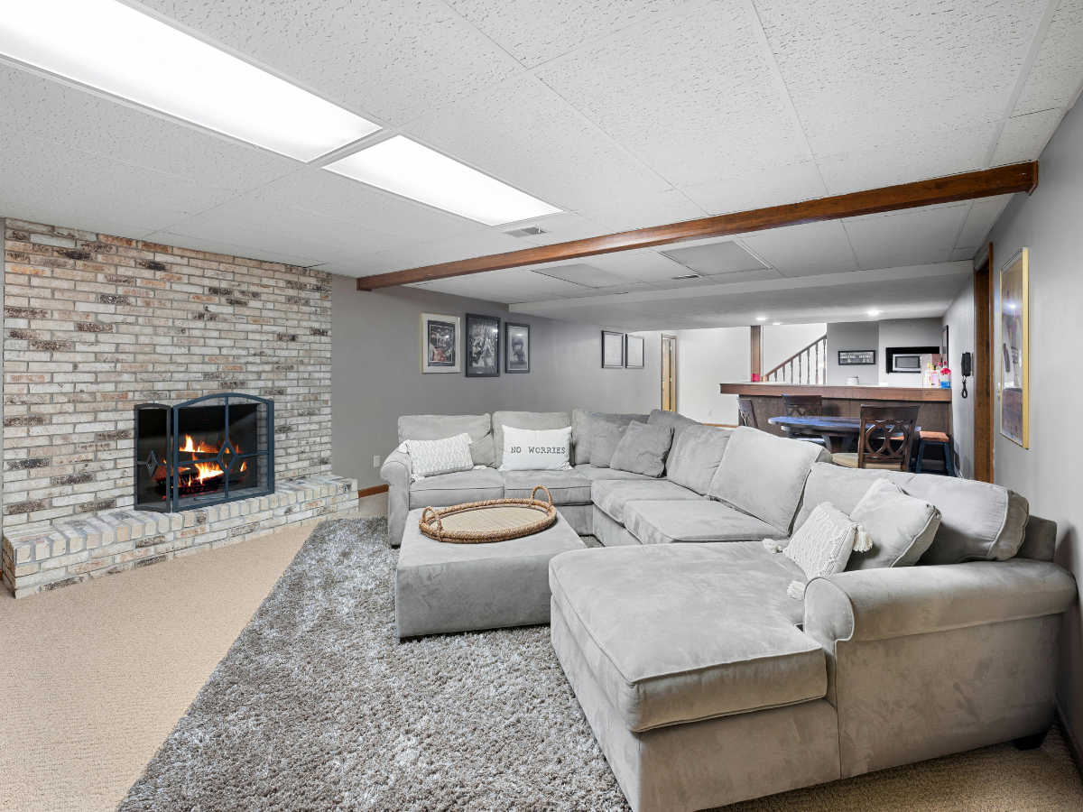Basement living room with gray sectional sofa, brick fireplace, carpeted floor, and wood beam ceiling; a tray is on the ottoman, and framed pictures hang on the walls—perfect inspiration for basement office design.