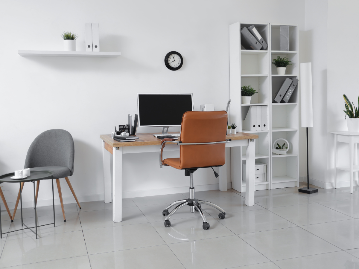 Modern basement office design featuring a desk, computer, brown chair, shelves with files, a round gray chair, a floor lamp, and plants against a white wall and tiled floor.