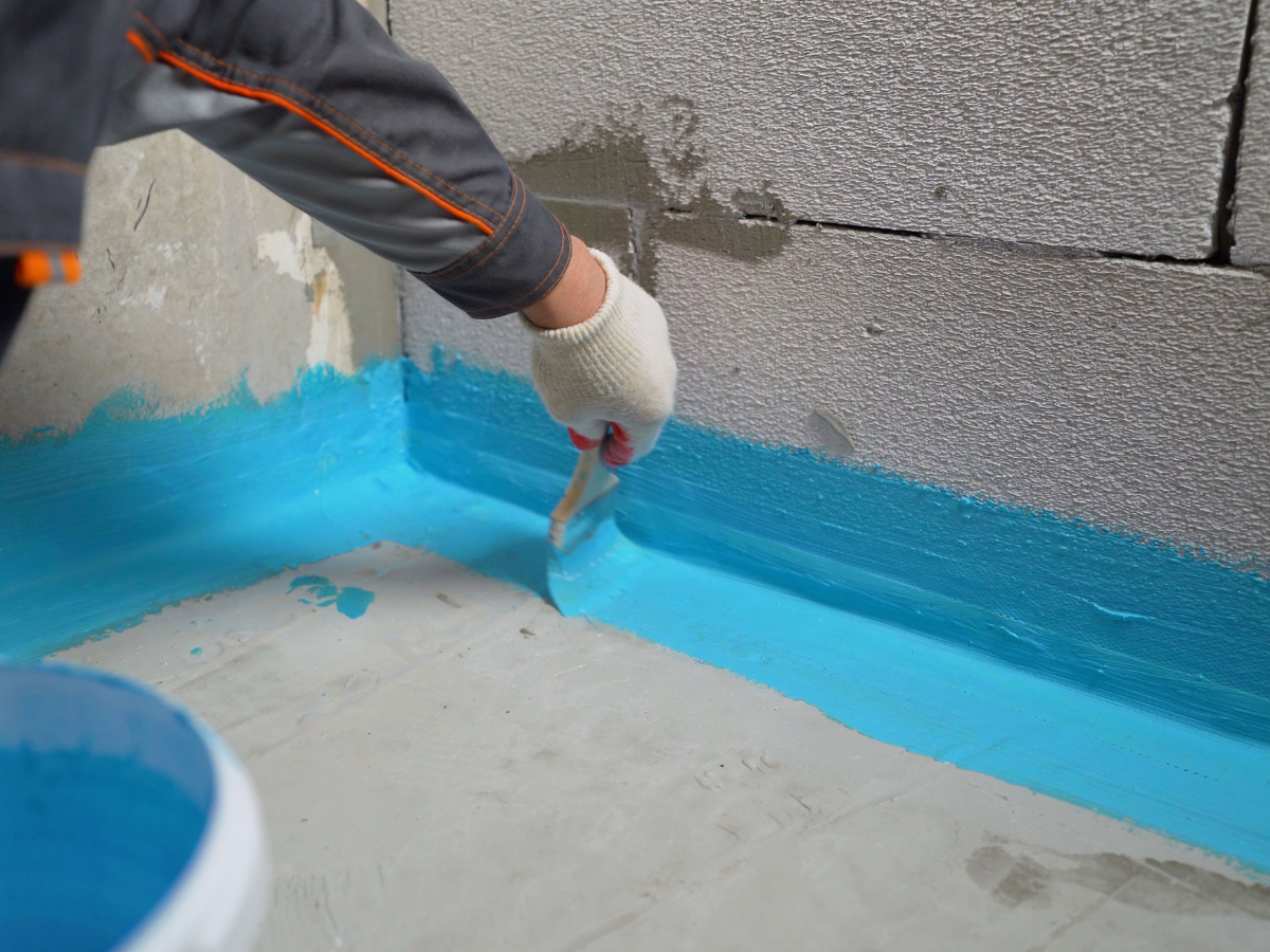 A person wearing gloves applies a blue basement waterproofing sealant to the corner of a concrete wall and floor using a brush.