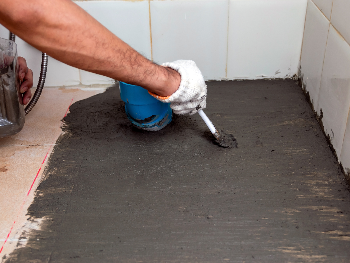 A person wearing a white glove spreads wet cement on a tiled surface using a small trowel, an essential step in basement waterproofing.
