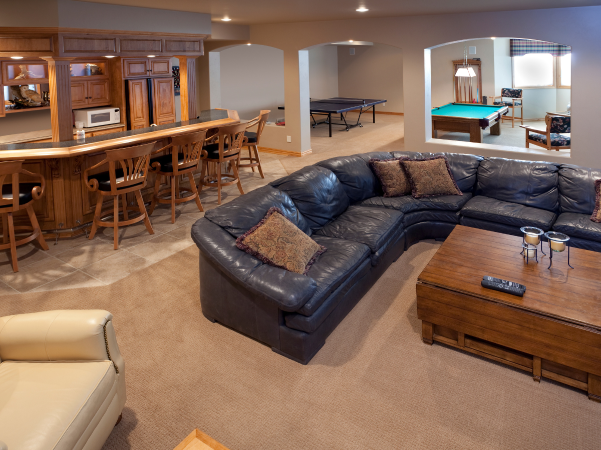 Spacious basement with a large sectional sofa, coffee table, bar area with stools, and game tables&mdash;including ping pong and pool&mdash;in the background. Recently updated with professional basement waterproofing for comfort and peace of mind.