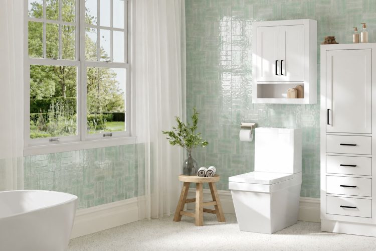Modern bathroom with a large window, a white bathtub, a square toilet, a wooden stool with plants and towels, and sleek ARIEL bathroom vanities and storage cabinets against green-tiled walls—perfect inspiration for your next office bathroom remodel.